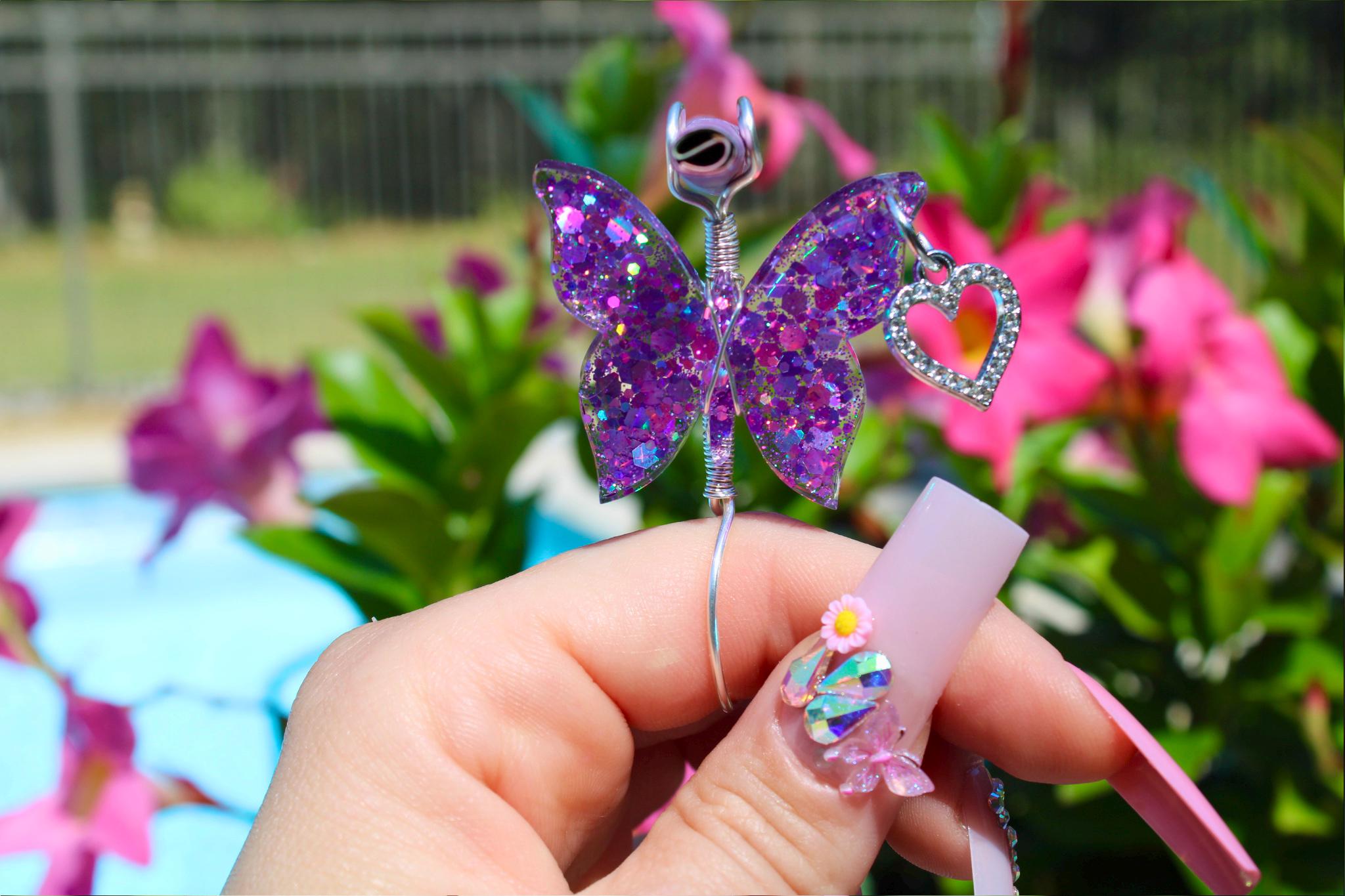 A hand with long, decorated nails holds a sparkly purple butterfly smoke ring featuring a heart charm, against a floral background.