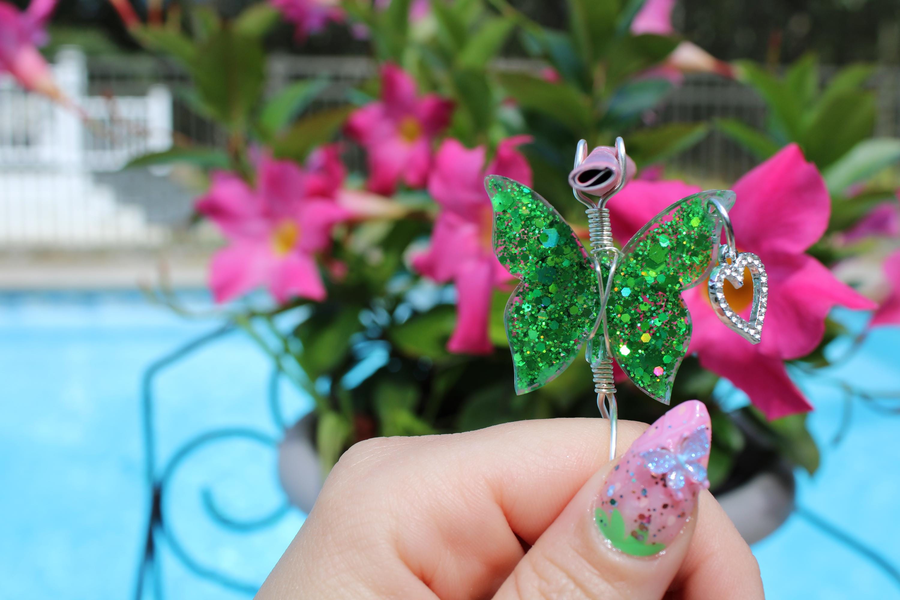 A hand with a butterfly-themed manicure holds a sparkly green butterfly smoke ring against a background of pink flowers and blue water.