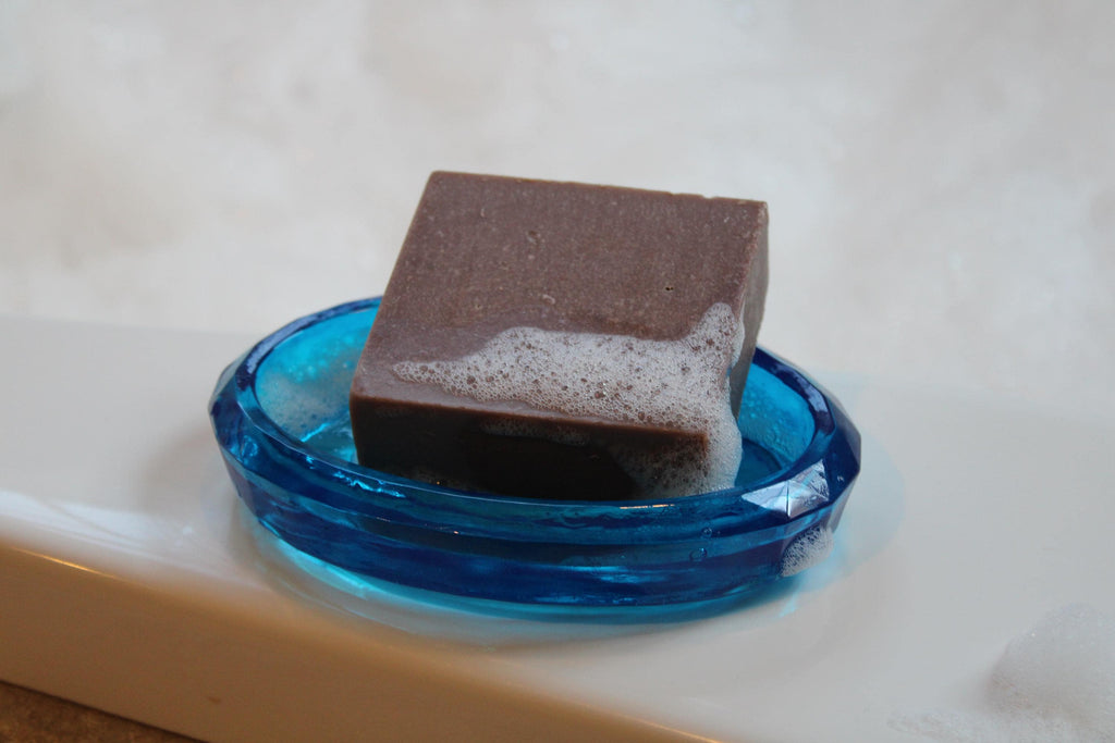 A square bar of natural brown soap sits in a decorative blue resin soap dish, showing soapy bubbles.