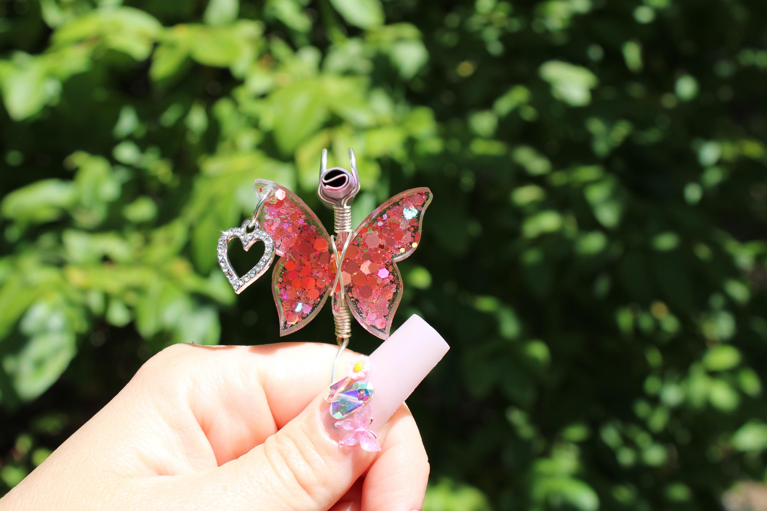 Woman's hand holds a sparkly pink butterfly smoke ring featuring a heart charm against a leafy green background.