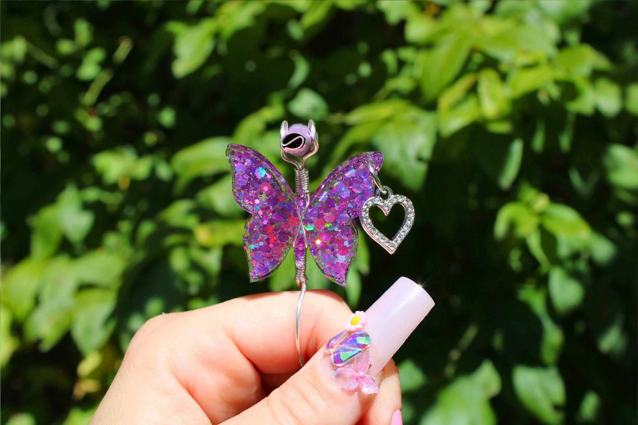 A manicured hand with butterfly nail art holds a sparkly purple butterfly smoke ring with a dangling heart charm against a leafy green background.