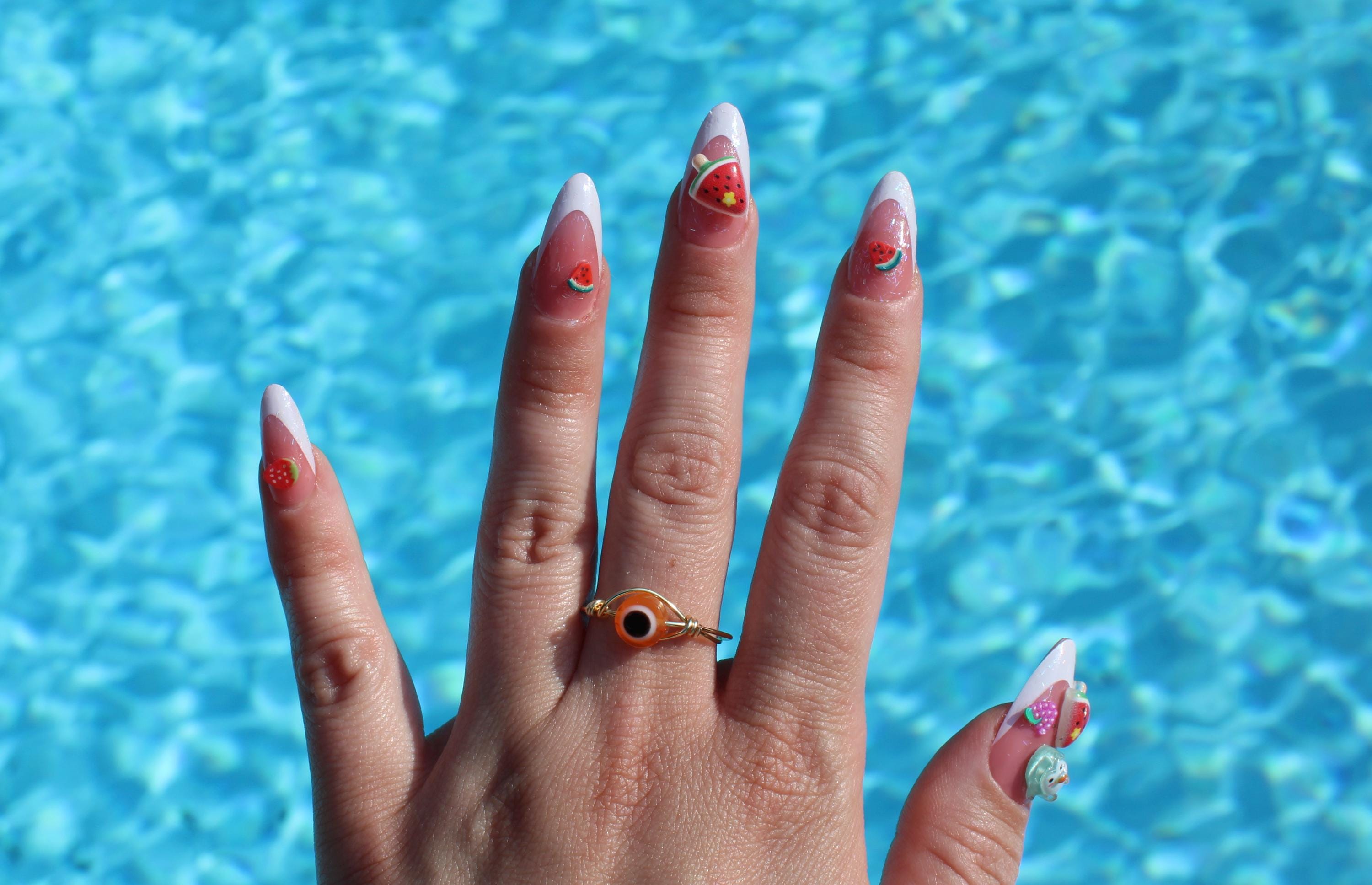 A hand models summer manicure art with fruit designs and an orange evil eye ring against sparkling blue pool water.