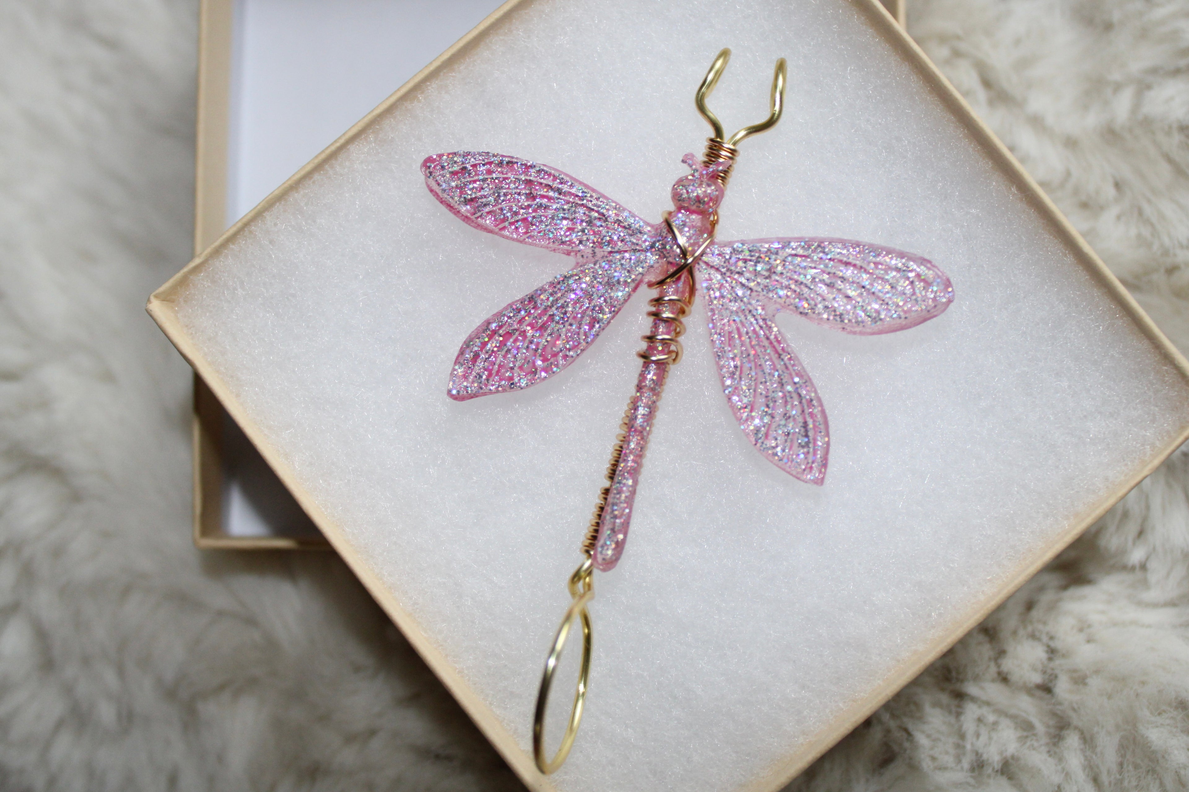 A sparkly pink dragonfly smoke ring rests in a white gift box, showcasing its intricate wire-wrapped design.