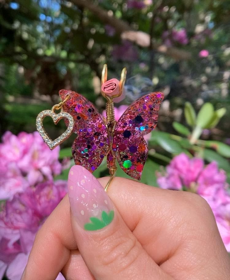 A hand with a strawberry-themed nail holds a handmade resin butterfly smoke ring with a dangling heart charm.