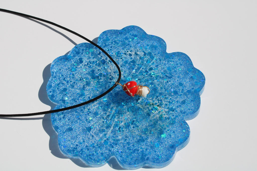 A blue, glittery resin flower coaster displays a red mushroom pendant necklace.