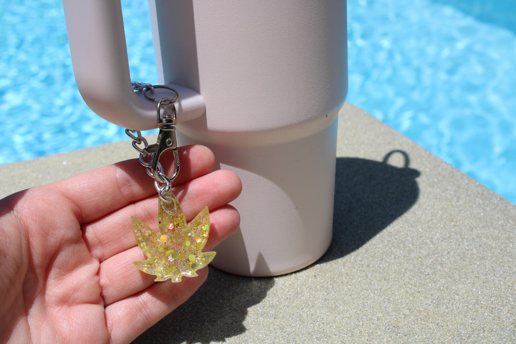 A hand displays a sparkling marijuana leaf keychain attached to a pale pink tumbler by a sunny pool.