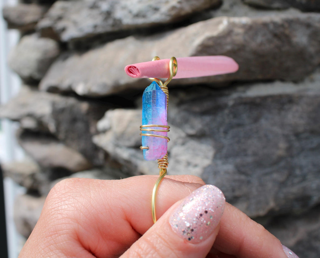 A hand wearing glittery pink nail polish displays a handcrafted gold wire smoke ring featuring a vibrant blue-and-pink crystal and a rolled pink paper detail.