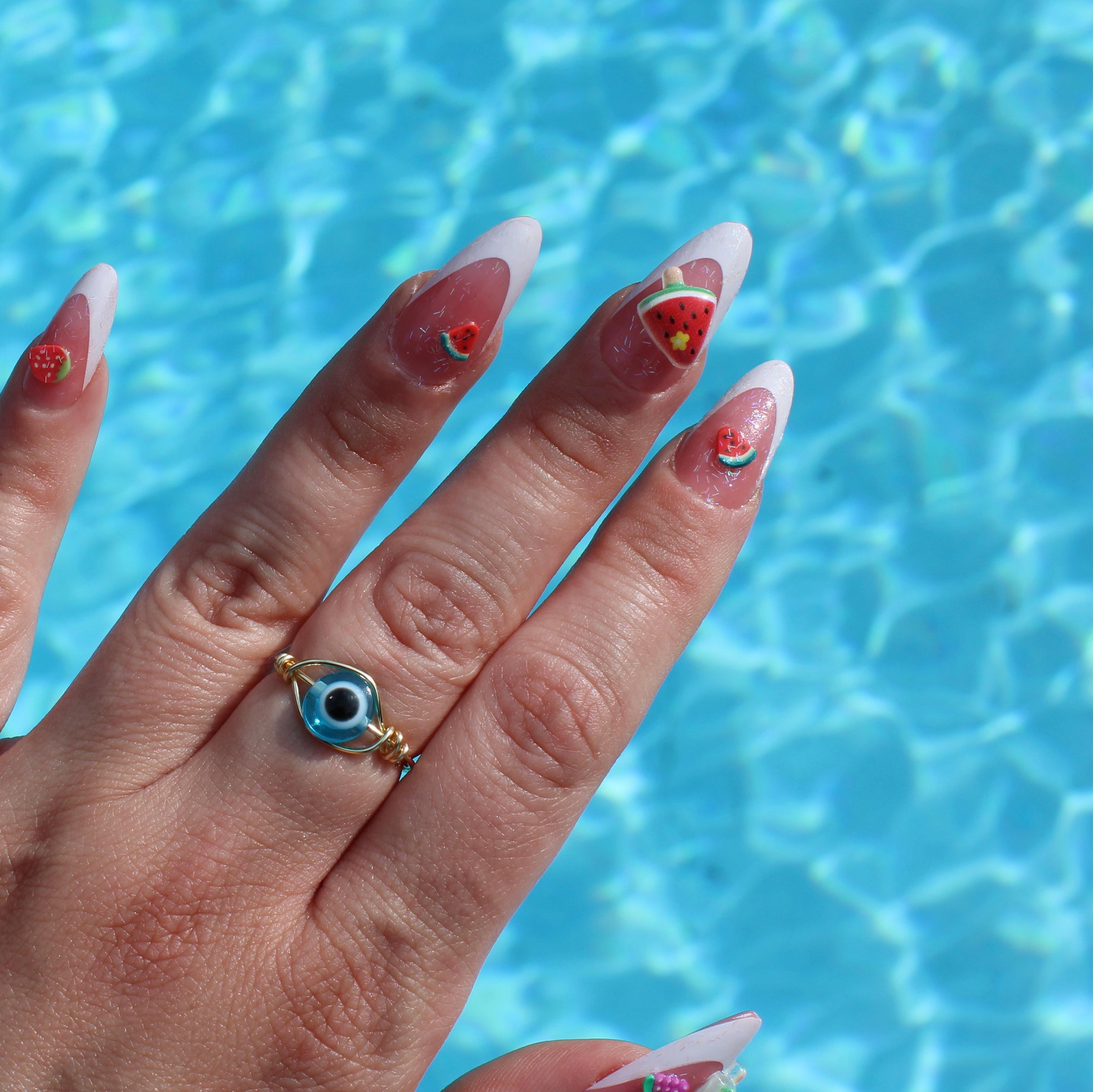 A hand wearing a light blue evil eye ring displays summer watermelon nail art over a shimmering blue pool.