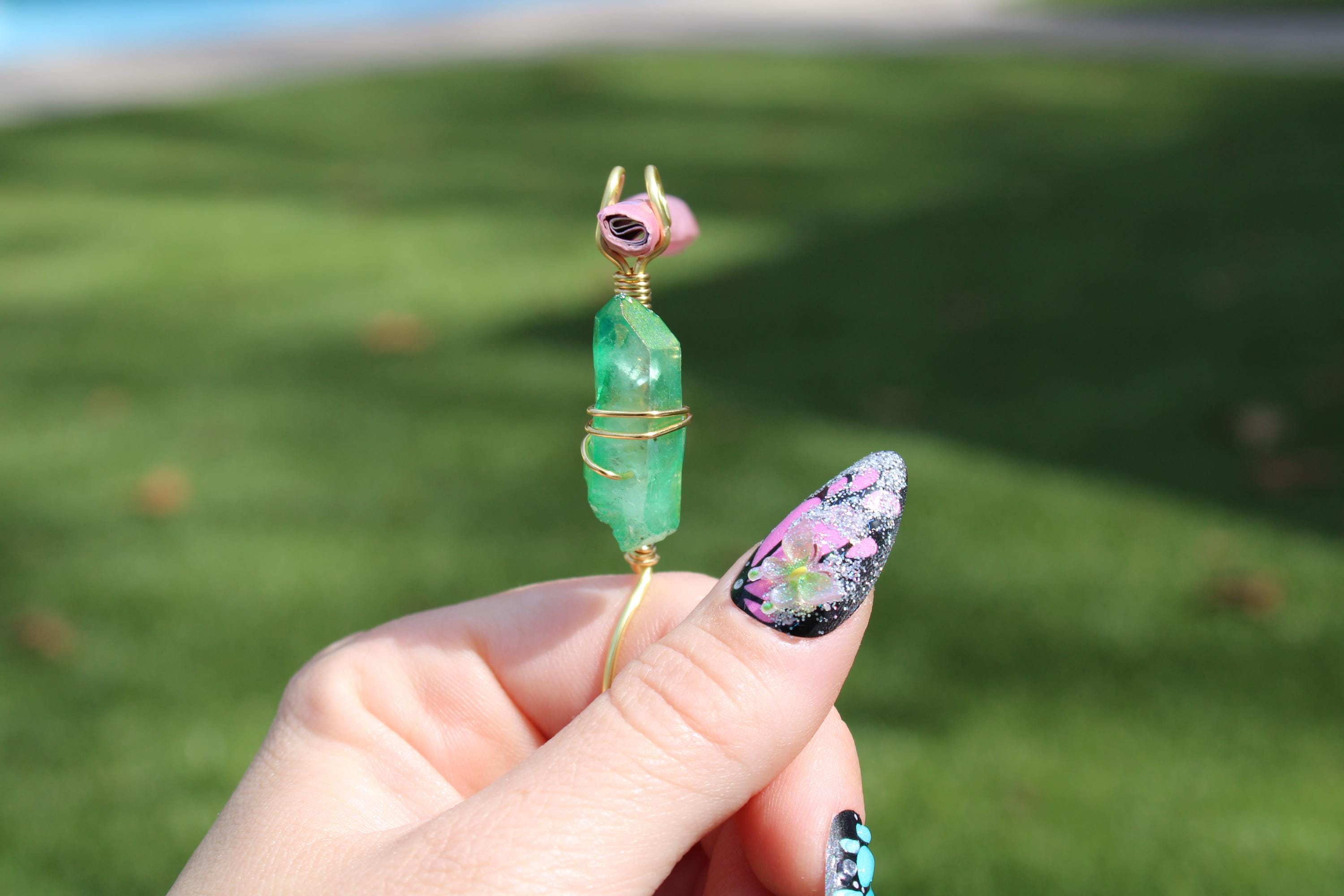 A hand with butterfly nail art holds a handmade green crystal smoke ring in an outdoor setting.