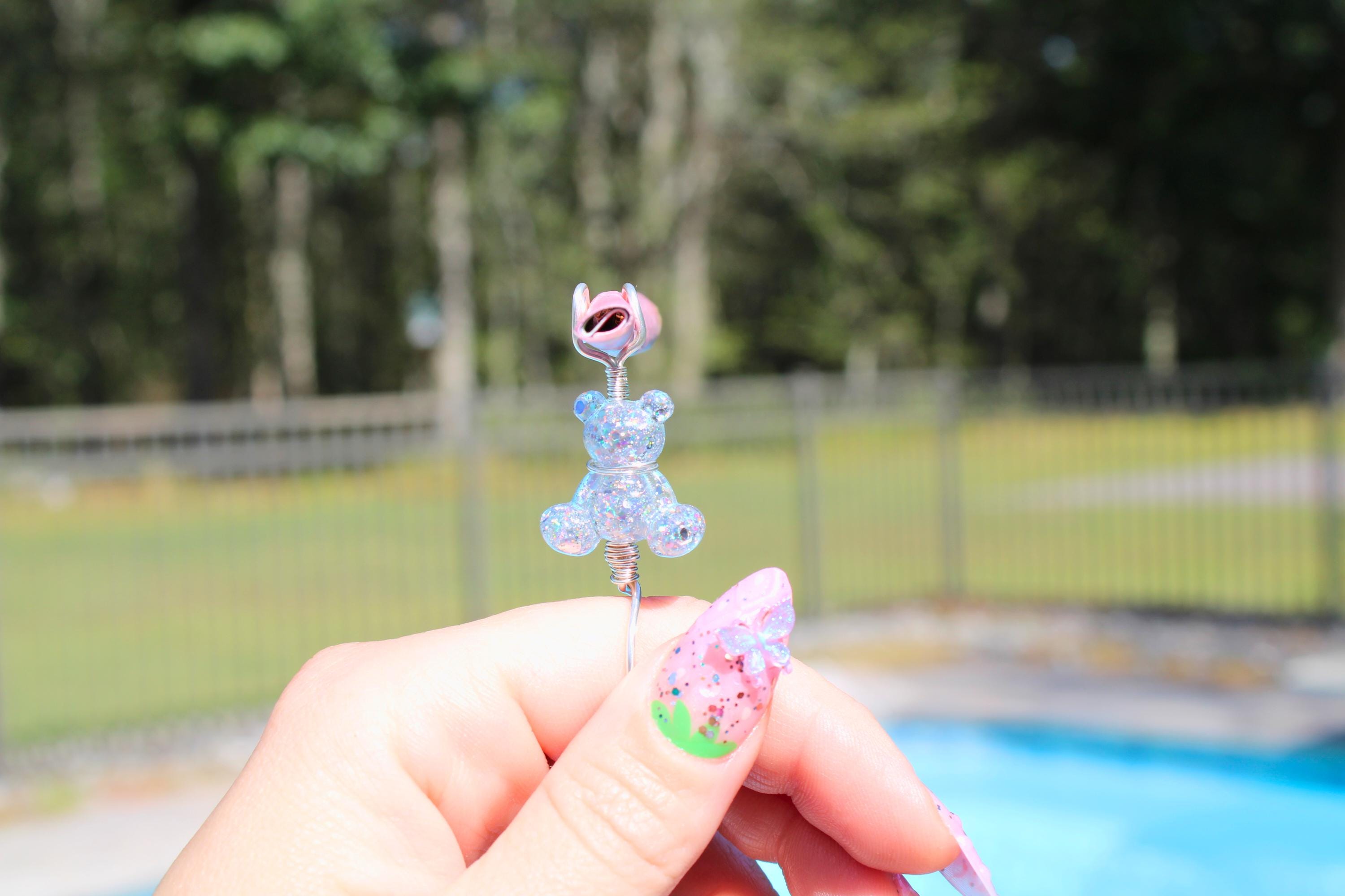 A hand with decorative nails holds a whimsical, handmade glitter teddy bear smoke ring against a blurred outdoor background.