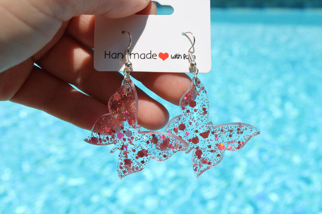 Butterfly-shaped earrings with pink speckles held by a hand against a pool background.