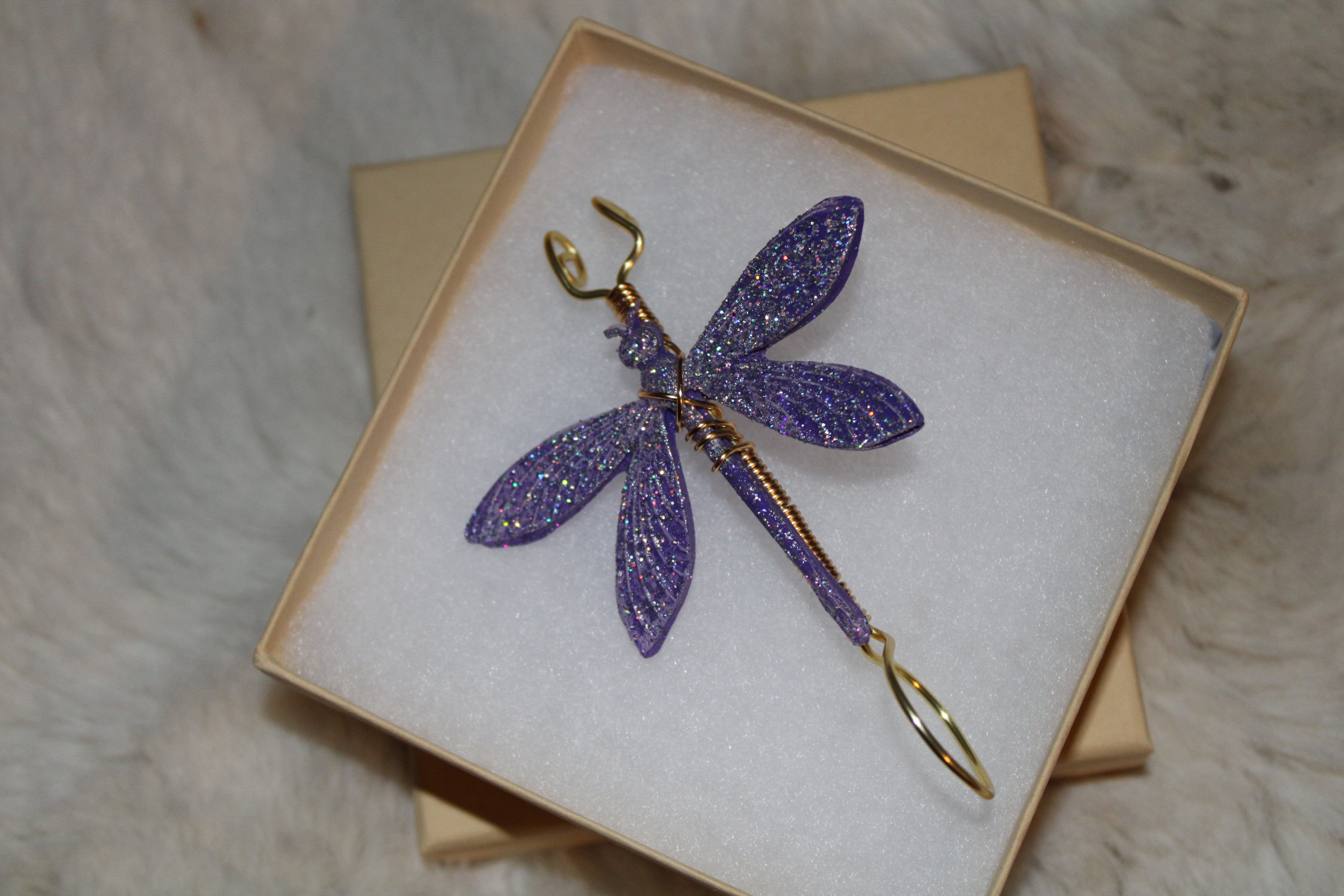 A glittery purple dragonfly smoke ring with gold wire accents rests inside a gift box.