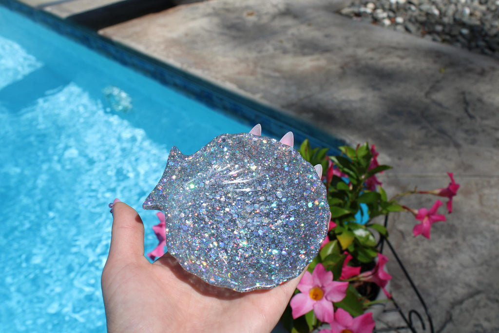 Hand holding a glittery silver shell by a pool with flowers in the background