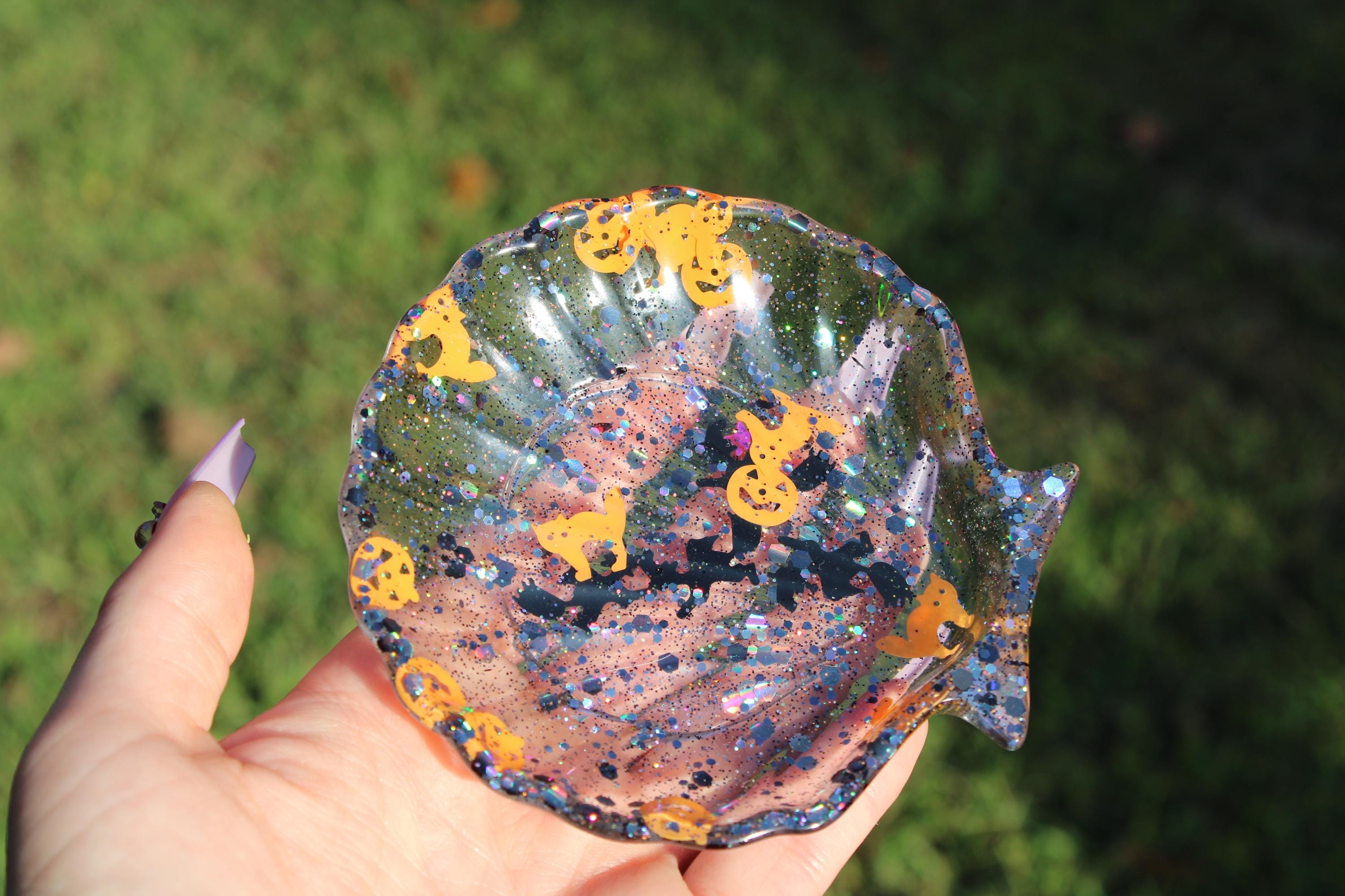 A hand holds a sparkling, clear resin Halloween trinket dish filled with glitter and spooky icons like pumpkins, bats, and ghosts.