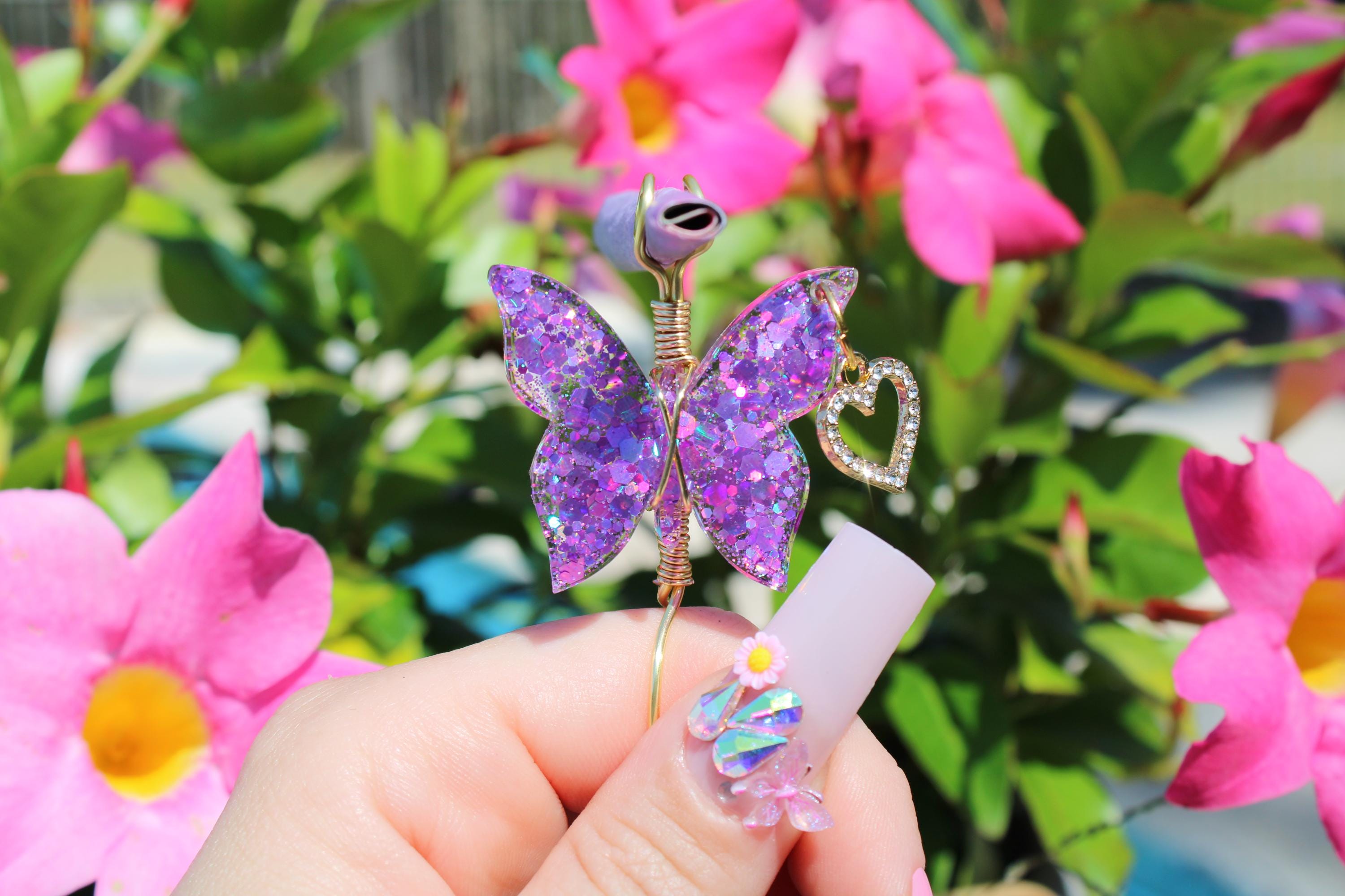 A hand with decorated acrylic nails holds a sparkly purple butterfly smoke ring against a backdrop of bright pink flowers.