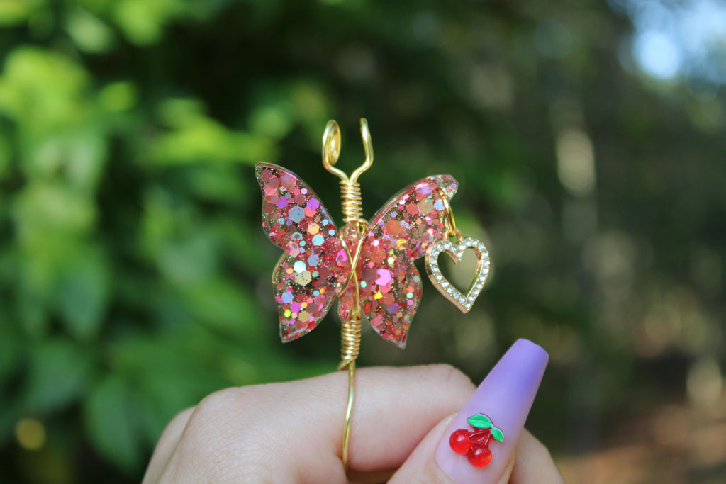 A hand with a lavender nail and cherry nail art displays a handmade glittery pink butterfly smoke ring featuring a gold heart charm.