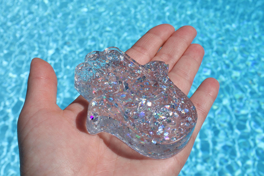 A hand holds a sparkling, translucent hamsa hand resin art over a bright blue swimming pool.