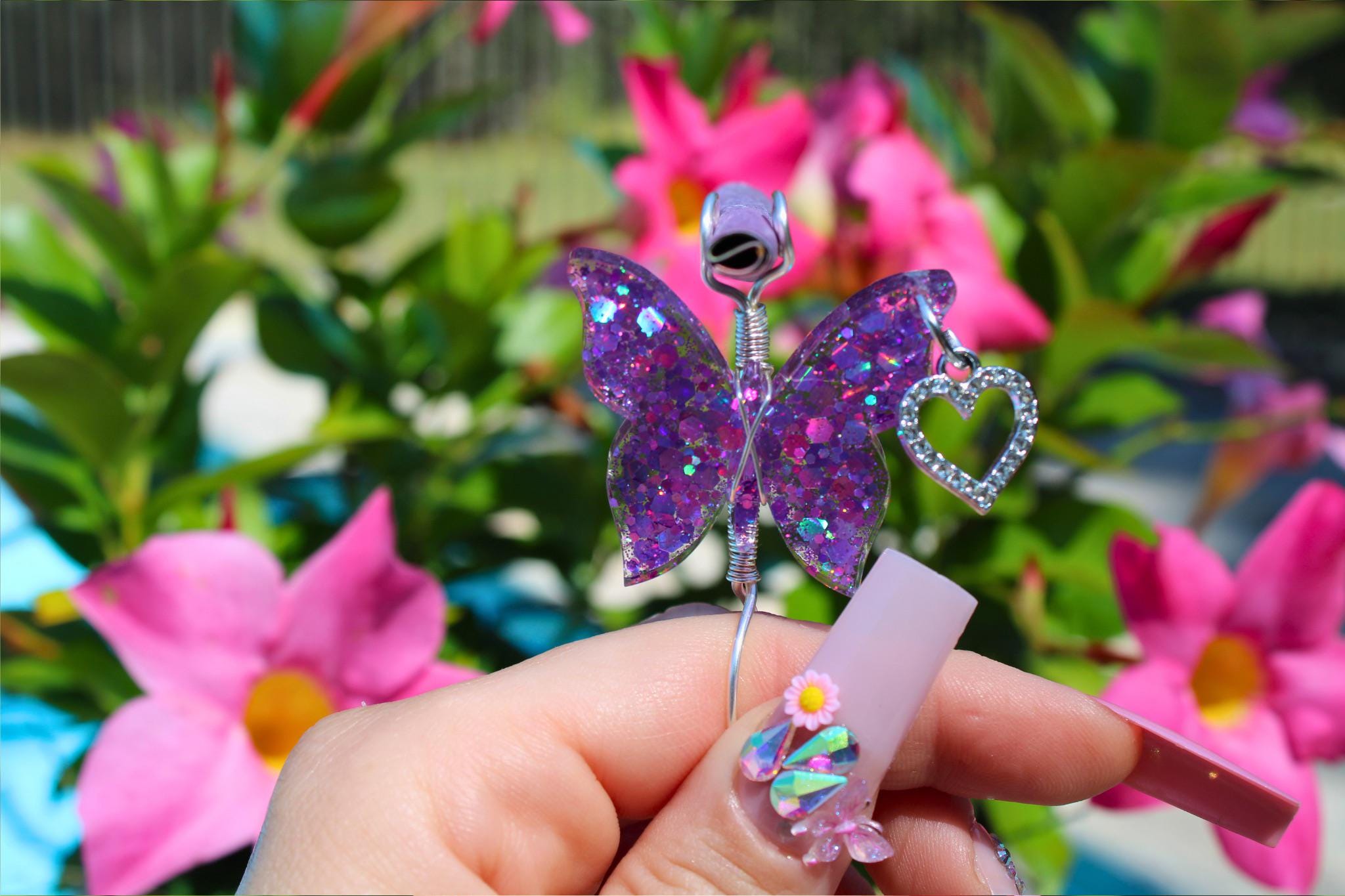 Woman shows off long acrylic nails adorned with 3D flower crystals while holding a glittery purple butterfly smoke ring holder against a backdrop of pink flowers.