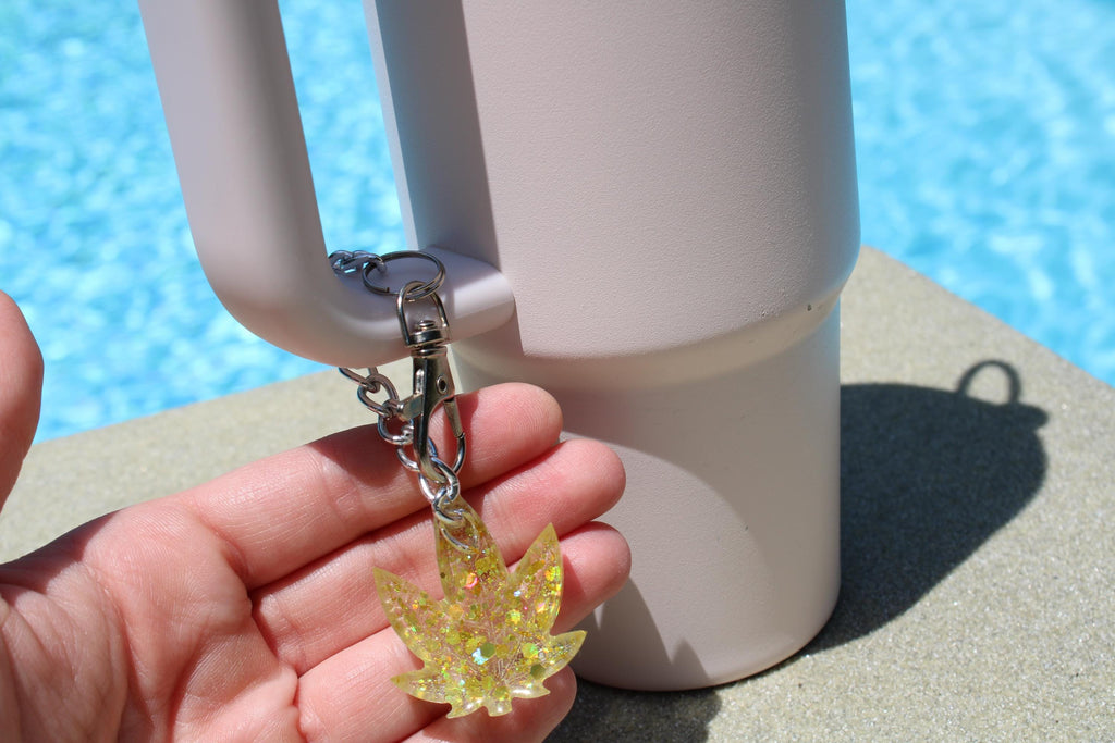 A hand displays a sparkly resin cannabis leaf keychain attached to a pink insulated tumbler near a pool.