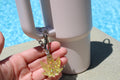 A hand displays a sparkly resin cannabis leaf keychain attached to a pink insulated tumbler near a pool.