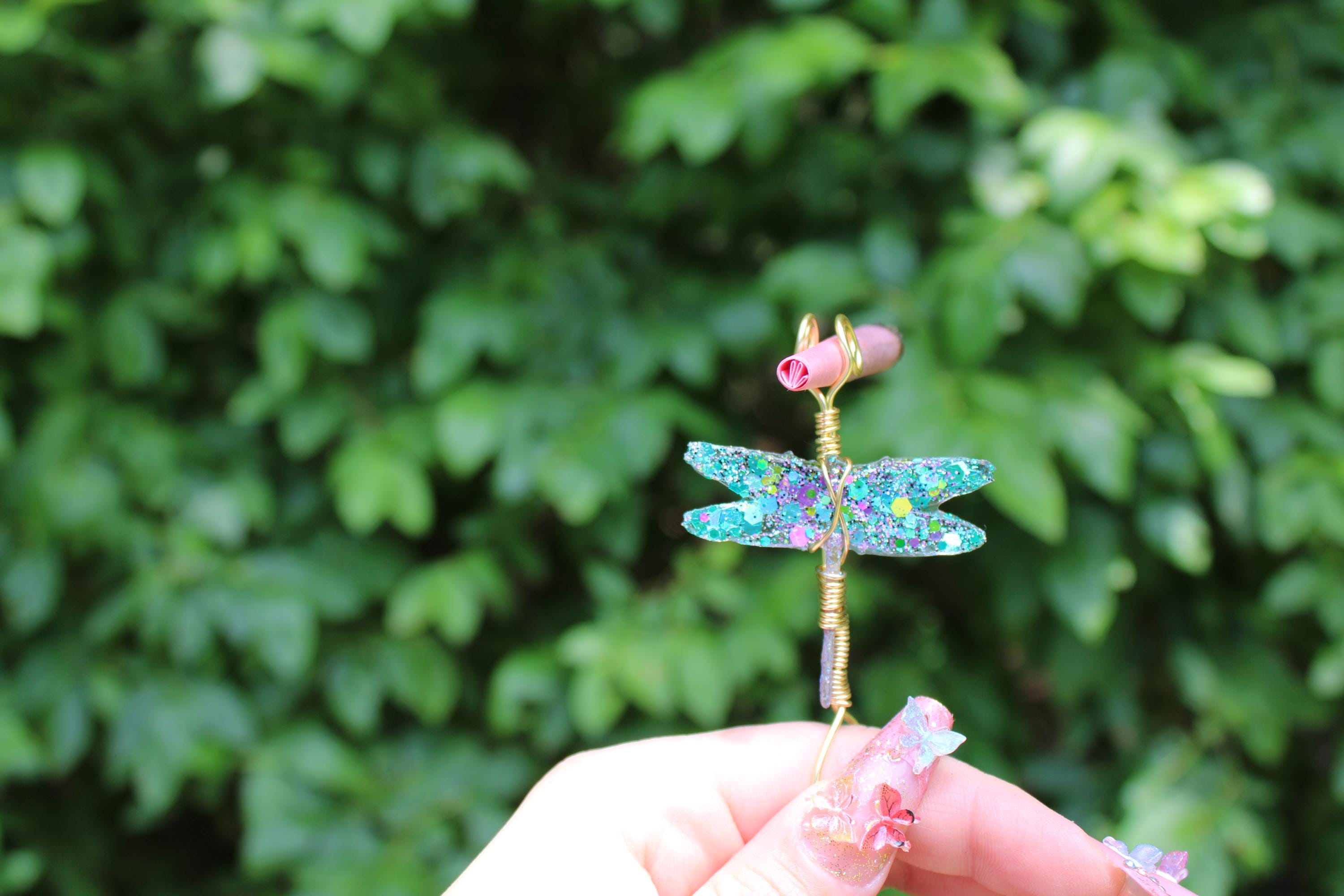 A manicured hand holds a whimsical, handmade wire dragonfly smoke ring featuring sparkly blue and purple wings against a green leafy background.