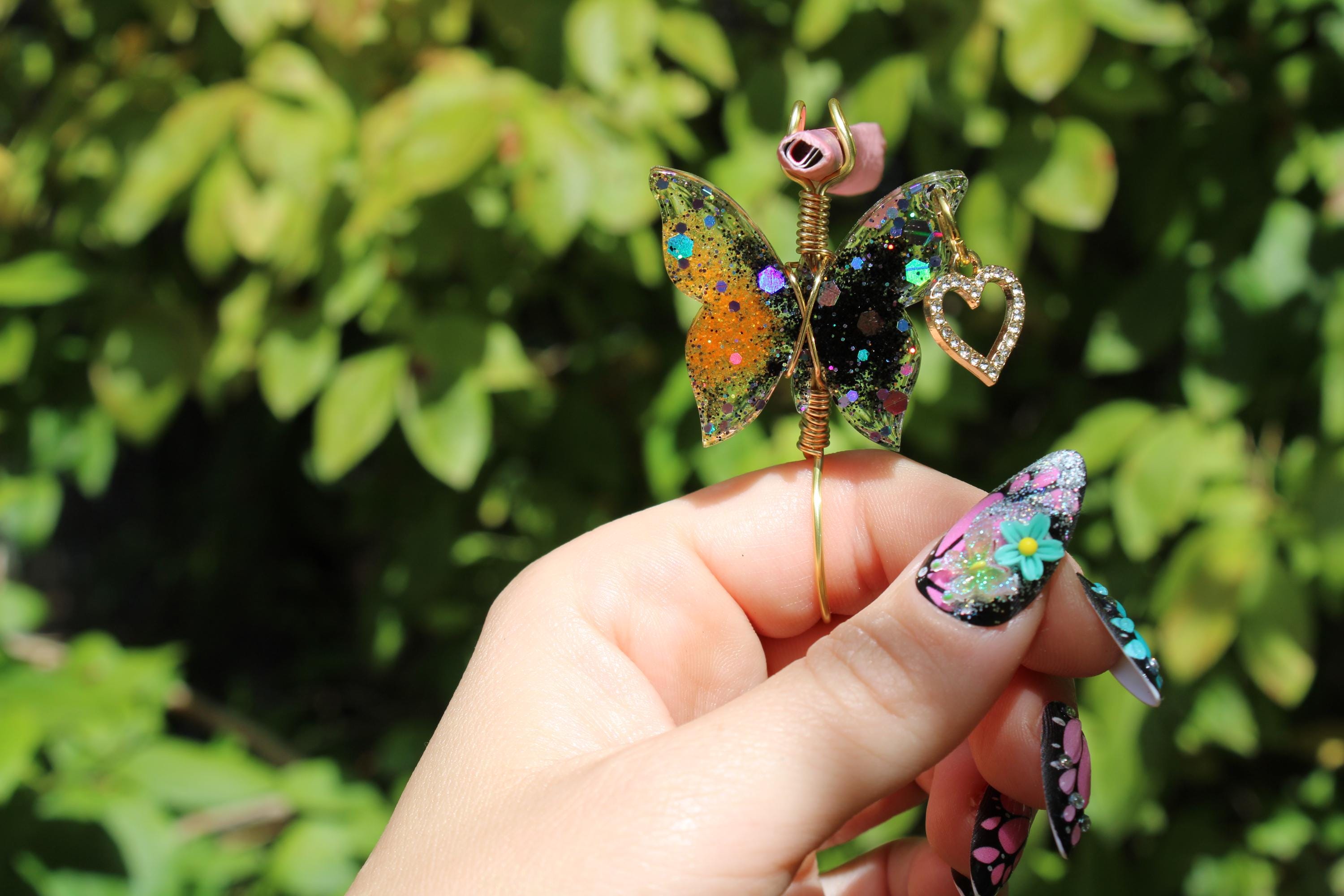 A hand with elaborate floral nail art displays a handcrafted butterfly smoke ring with a rose and heart charm against a leafy background.