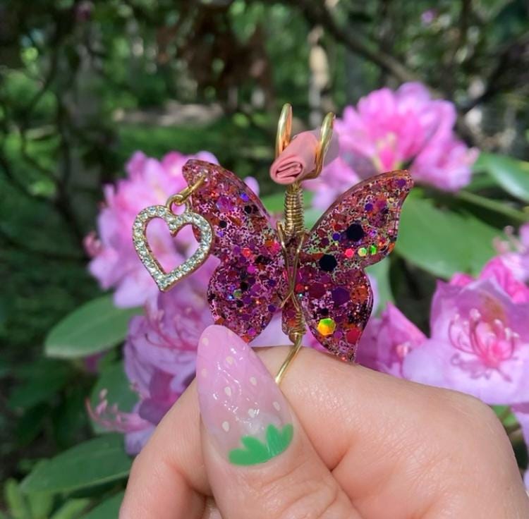 A hand with a strawberry-themed nail holds a handmade glittery butterfly smoke ring with a heart charm, set against a blooming pink flower bush.
