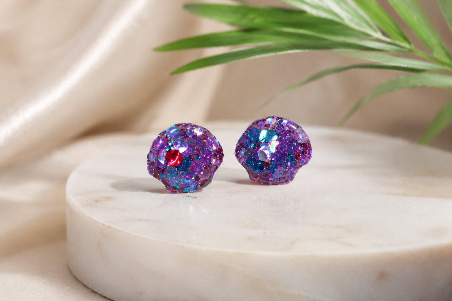 A pair of shell stud earrings covered in iridescent purple, blue and pink sequins displayed on a round white marble slab with a green leaf in the background.