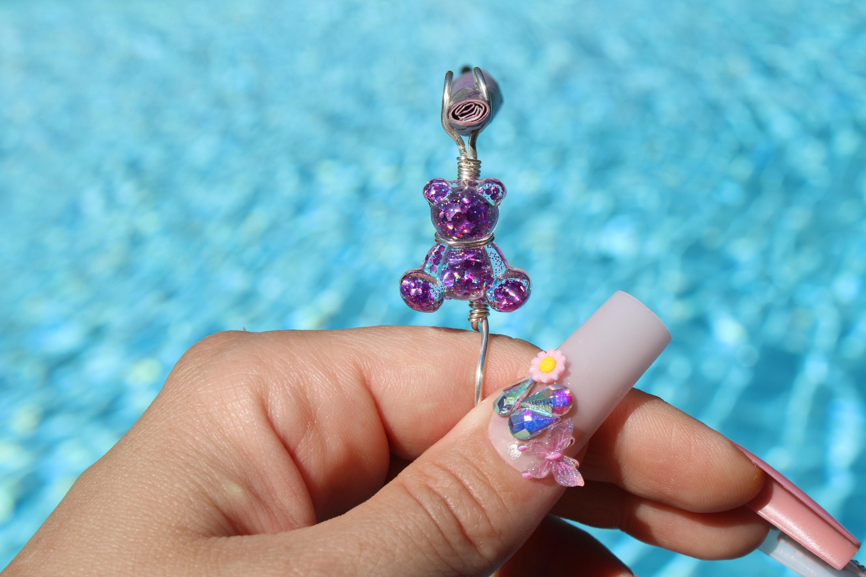 A hand displays a long, pastel pink acrylic nail art holding a purple sequin teddy bear smoke ring, set against a bright blue pool background.
