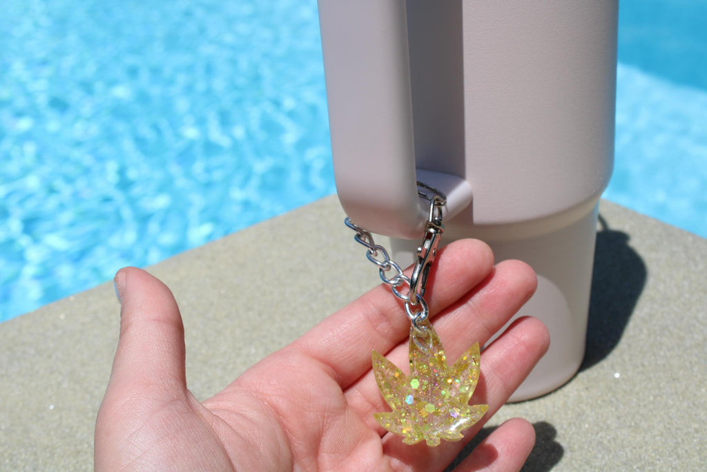 A hand holds a sparkly yellow cannabis leaf keychain attached to a tumbler cup by the pool.
