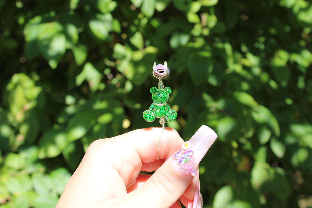 A hand with decorated nails holds a whimsical, green glitter teddy bear smoke ring clip against a leafy green background.