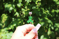 A hand with decorated nails holds a whimsical, green glitter teddy bear smoke ring clip against a leafy green background.