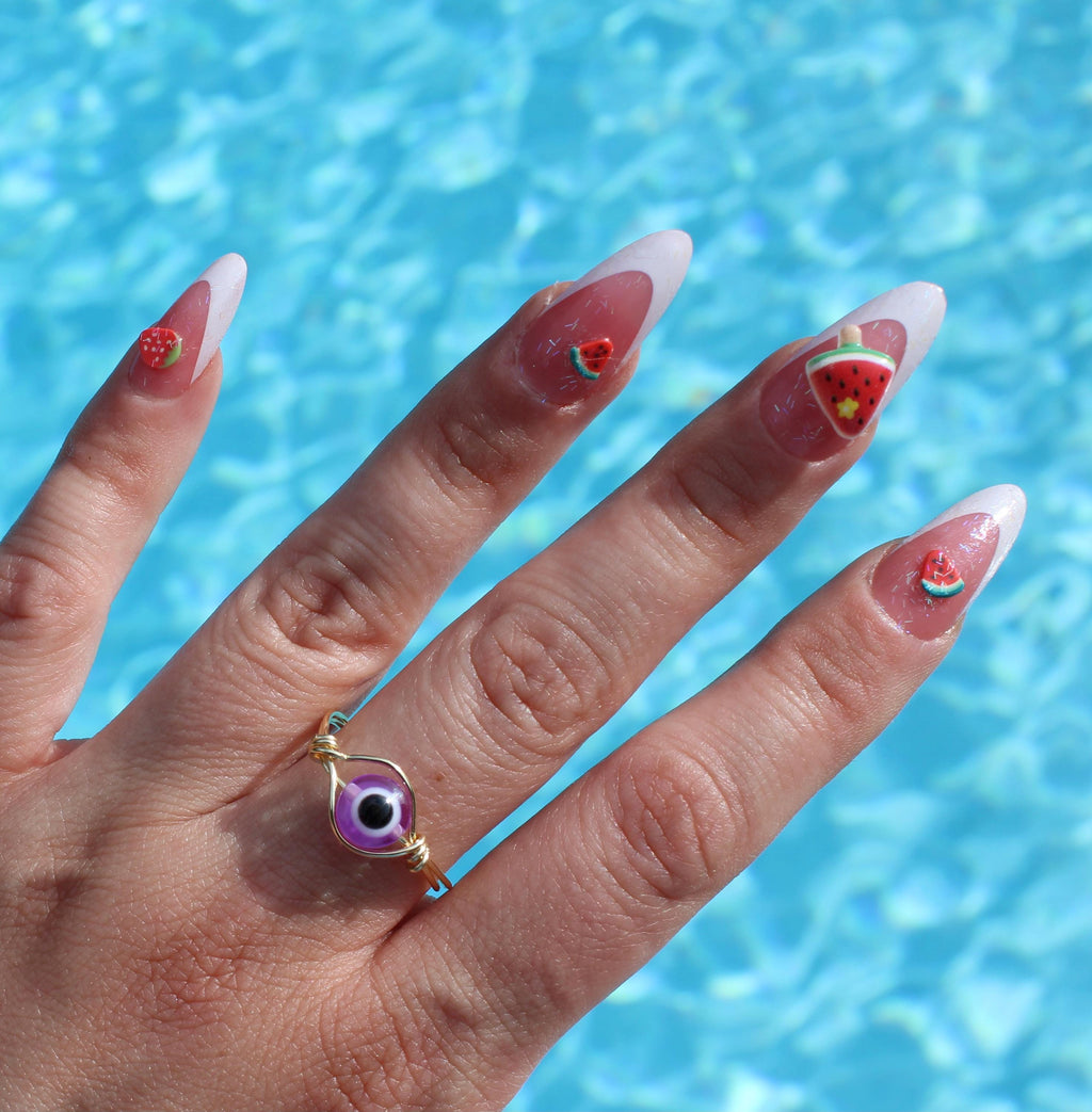 A hand wearing an purple evil eye ring displays long, summer-themed nails with watermelon and strawberry designs against a bright blue pool background.