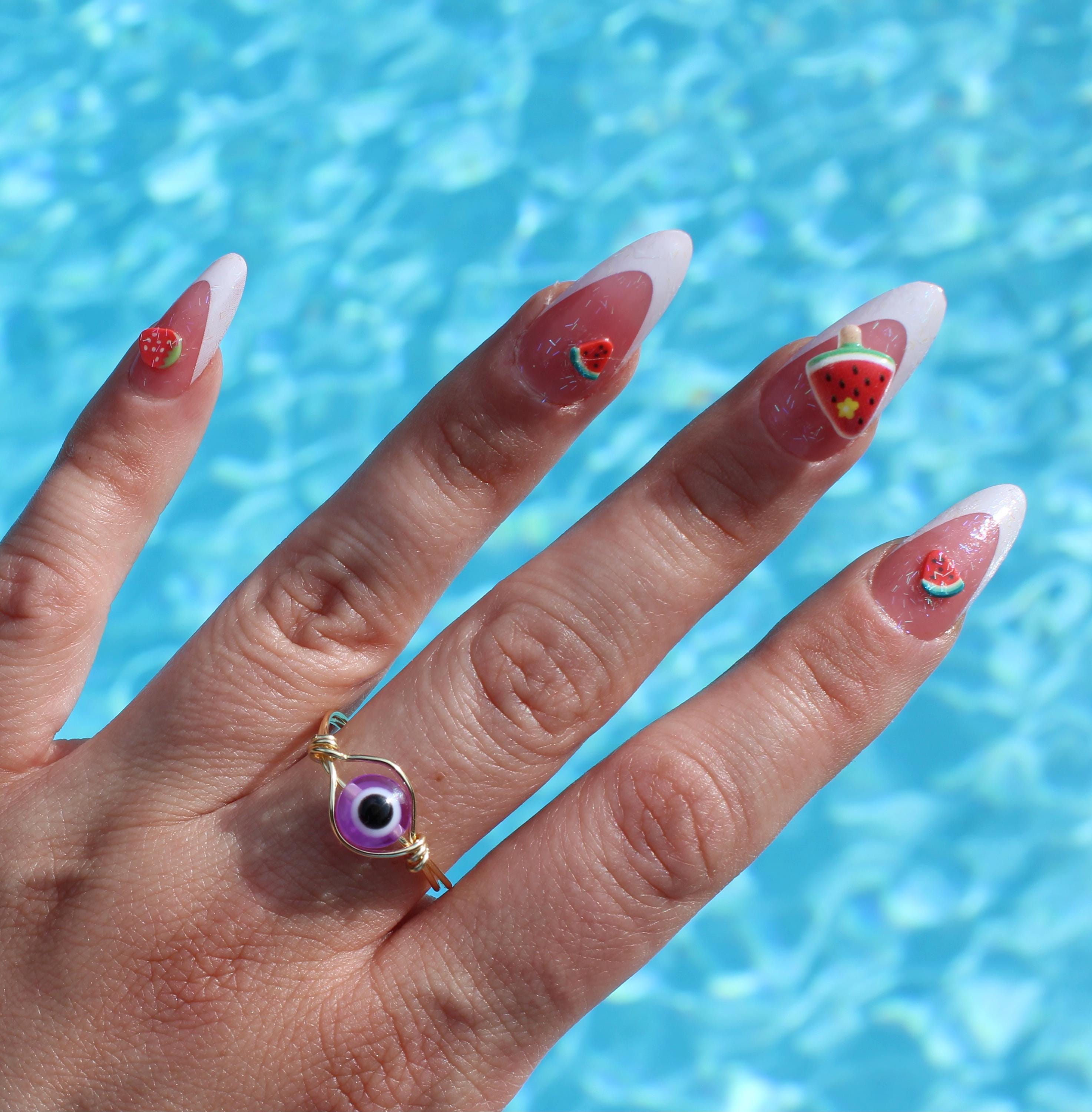 A hand wearing an purple evil eye ring displays long, summer-themed nails with watermelon and strawberry designs against a bright blue pool background.