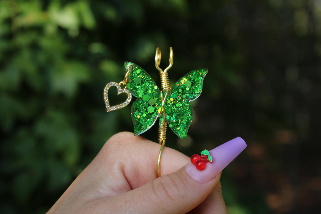 A hand models a gold colored wire green butterfly smoke ring featuring green glitter wings and a heart charm, showcasing a trendy lavender manicure adorned with a cherry design.