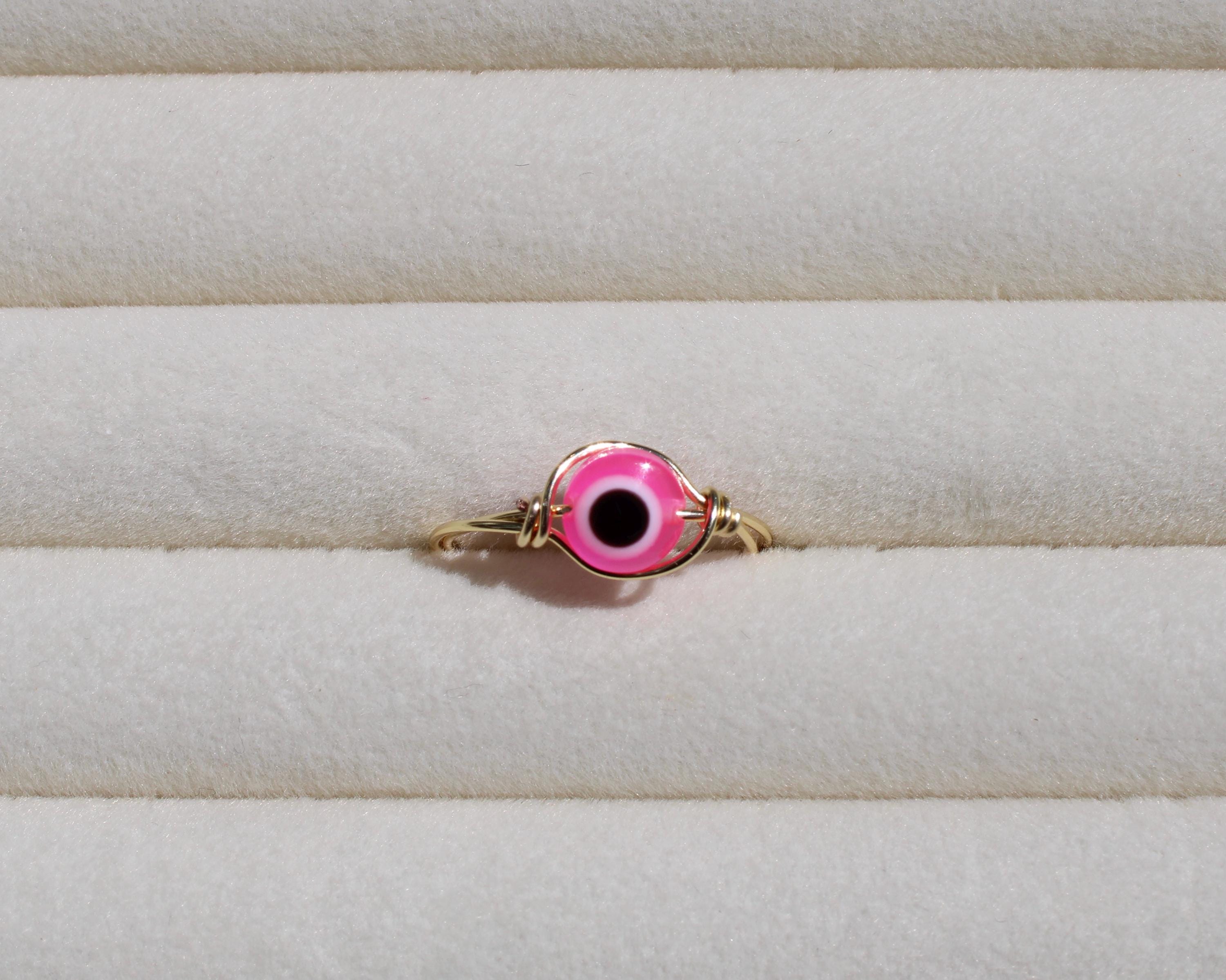 A gold wire ring displays a pink evil eye bead on a textured cream surface.