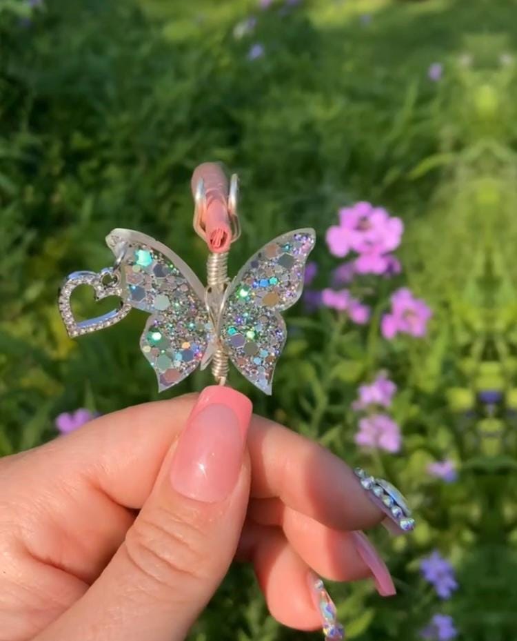 A hand with stylish, pink-toned nails holds a sparkly butterfly smoke ring against a blurred, flowery green backdrop.