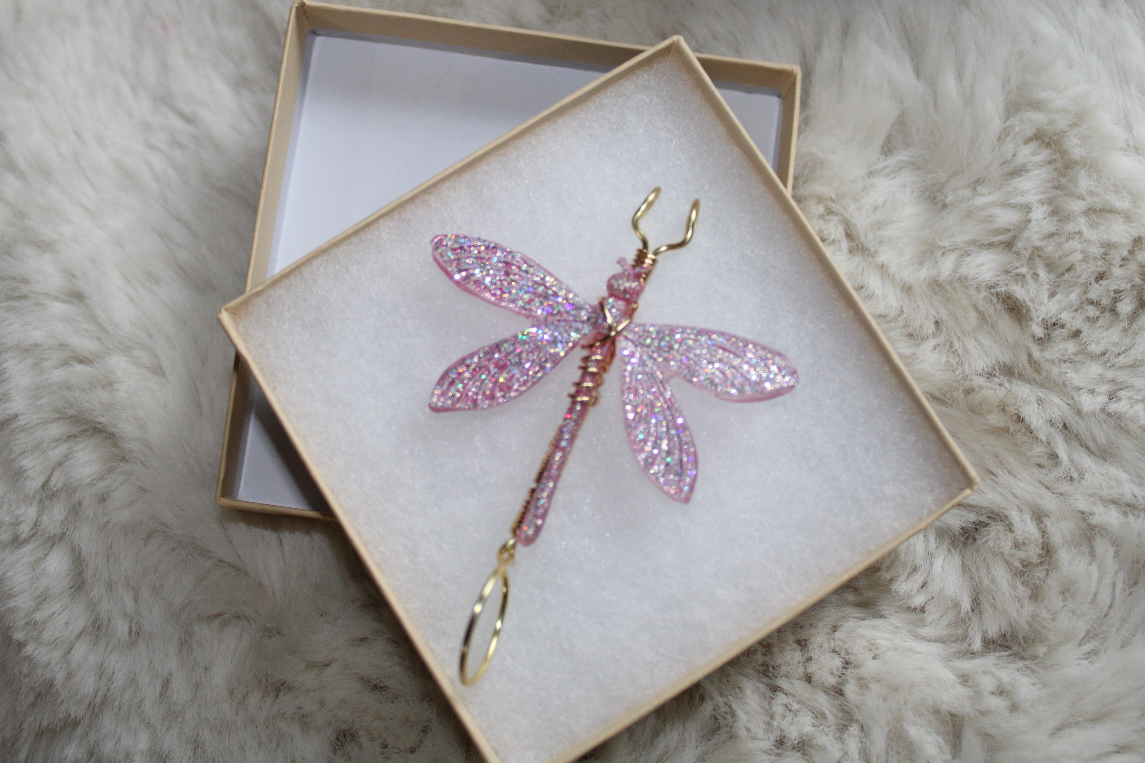 A handmade pink glitter dragonfly smoke ring rests inside a beige gift box on a fluffy white surface.