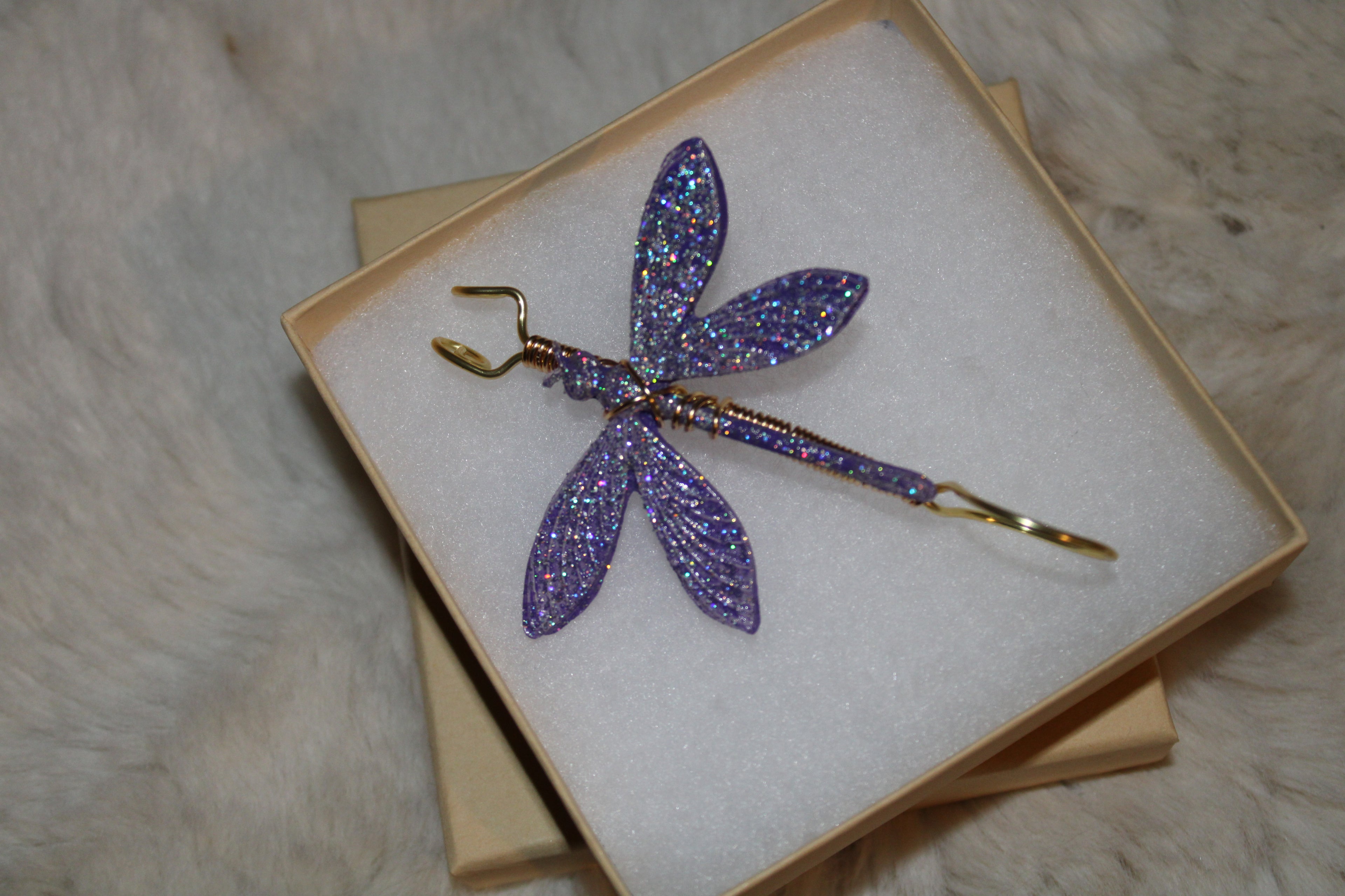 A sparkly purple dragonfly smoke ring with gold wire body sits nestled in a gift box.