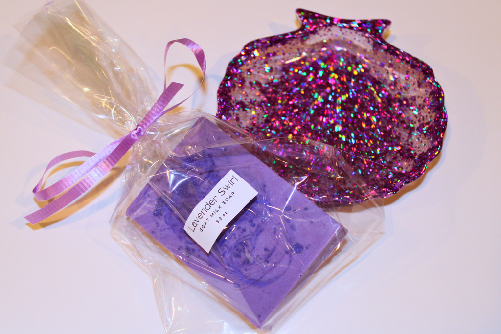 A wrapped lavender swirl goat milk soap sits next to a sparkling purple resin seashell.