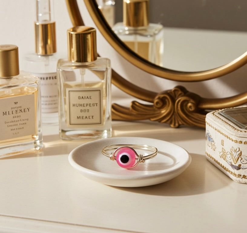 Pink evil eye ring rests on a white dish amongst perfume bottles and a decorative mirror on a vanity.