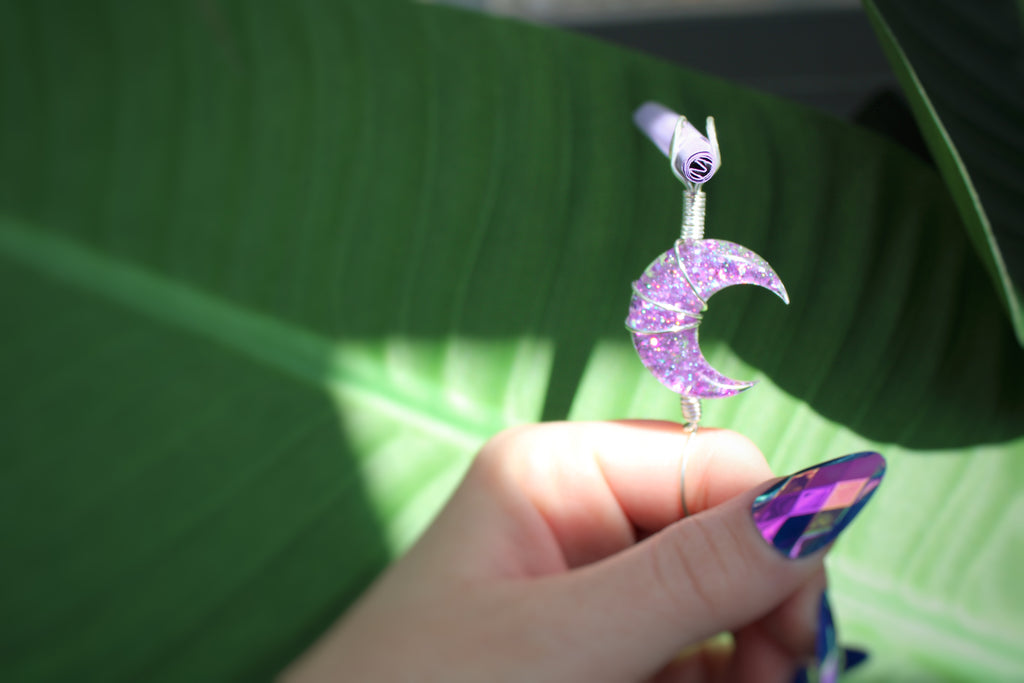 Hand holding a purple glittery crescent moon smoke ring against a green leaf background
