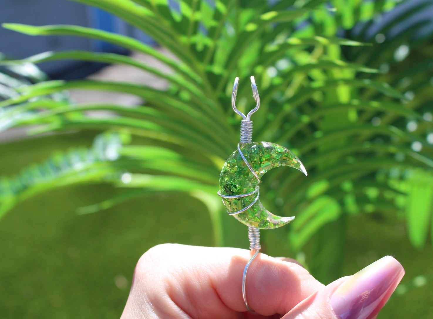 Hand holding a green wire-wrapped crescent moon smoke ring with a blurred green plant background
