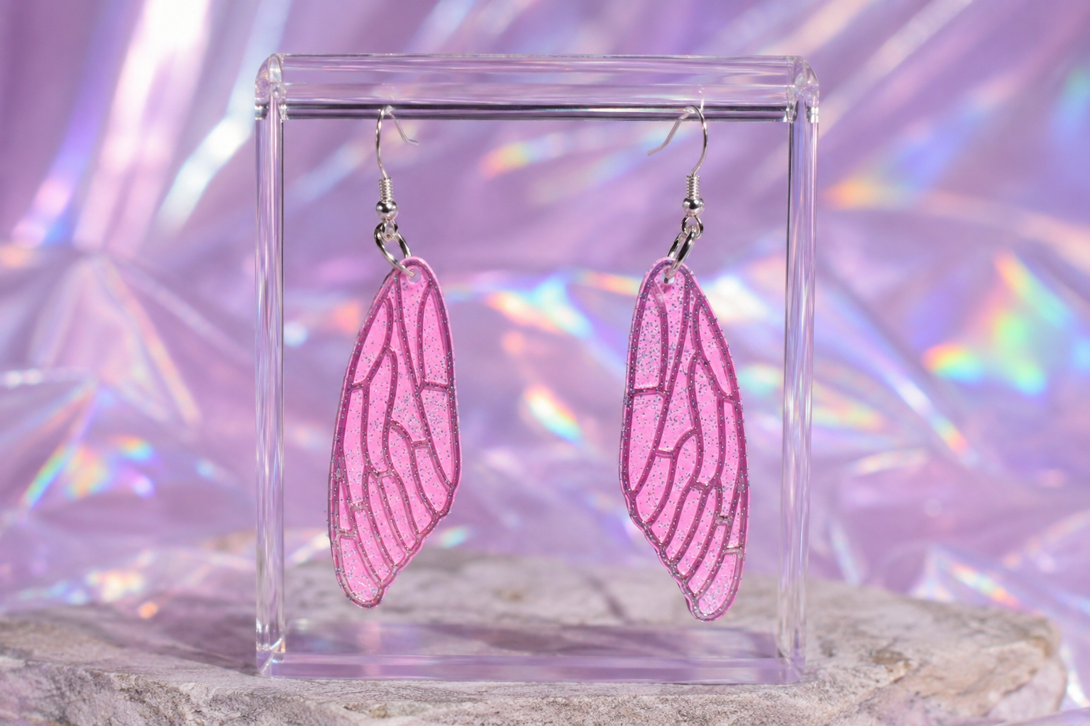 Pink wing-shaped earrings on a clear stand with a colorful, iridescent background.