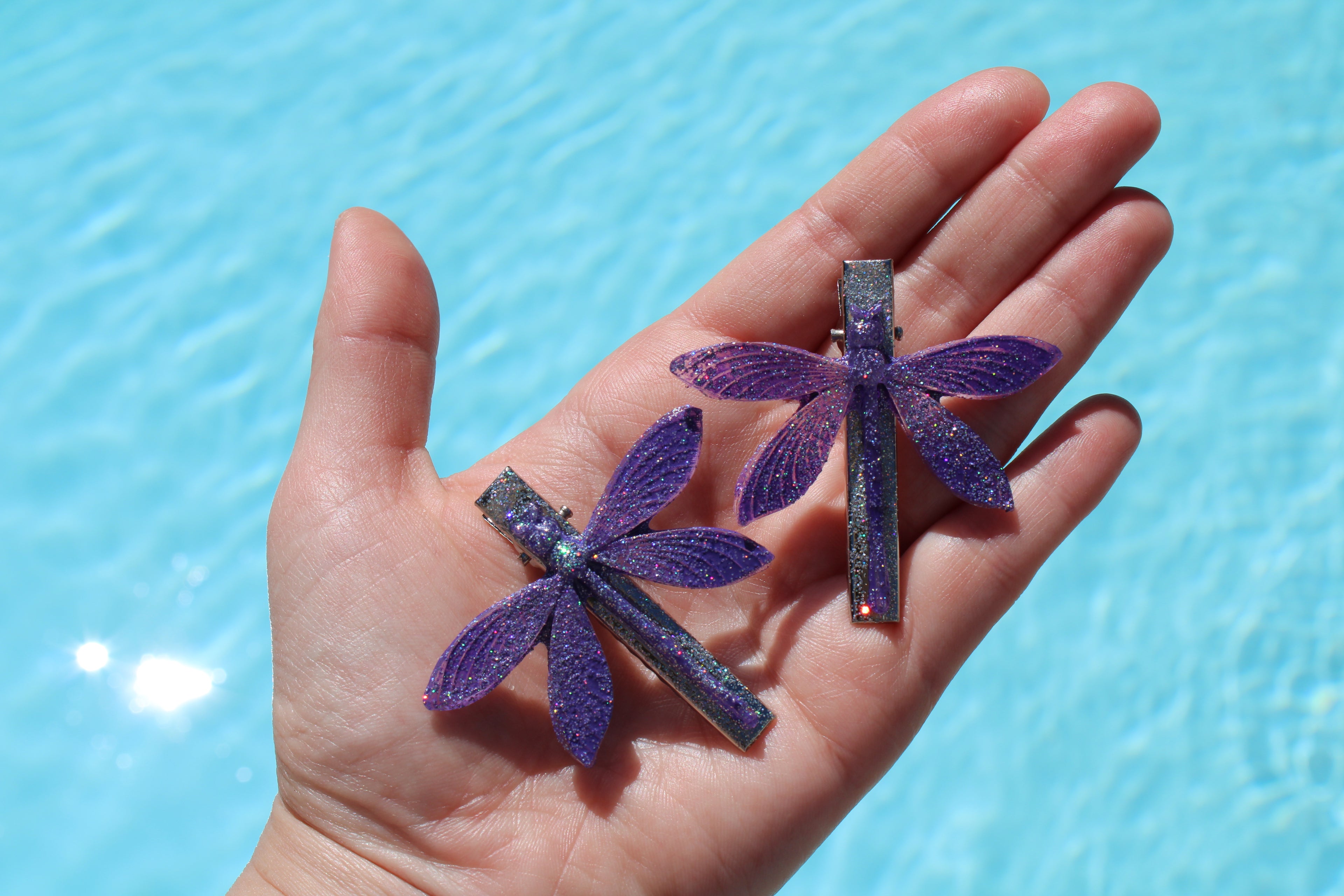Hand holding two purple dragonfly-shaped hair clips against a blue water background