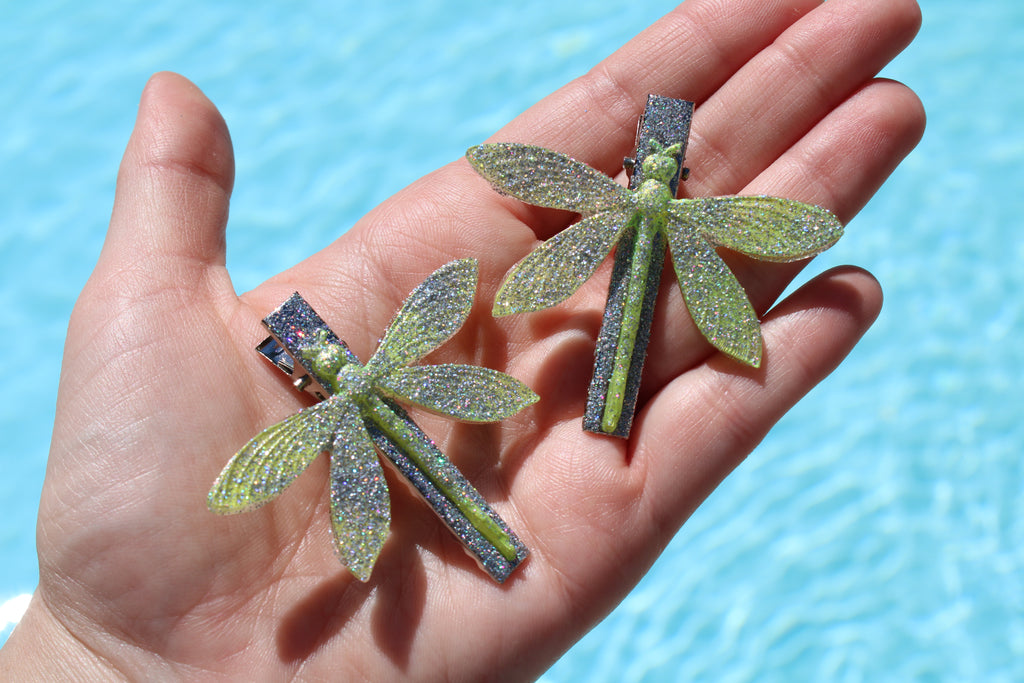 Two glittery dragonfly-shaped hair clips held in a hand with a blue background