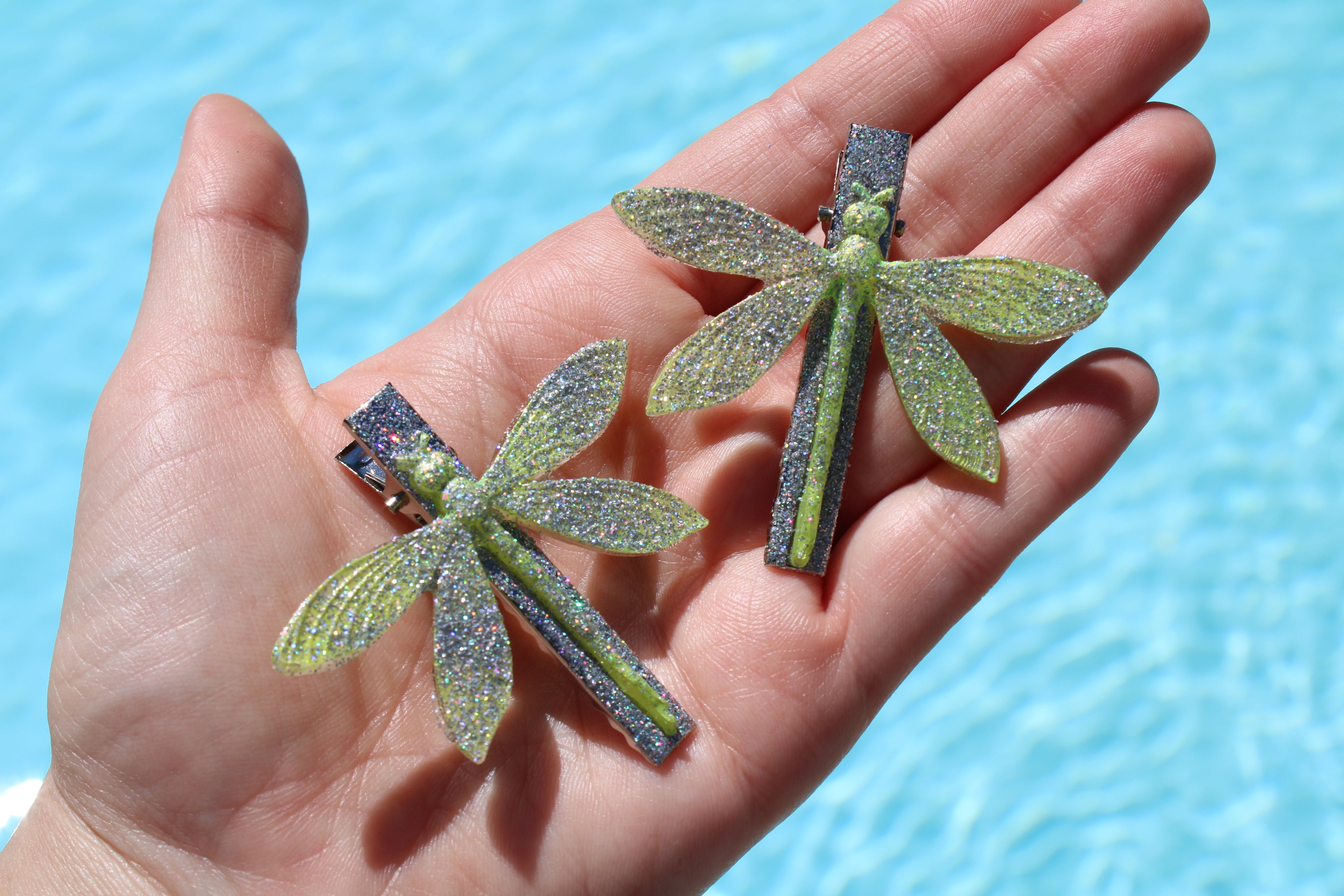 Two glittery dragonfly-shaped hair clips held in a hand with a blue background