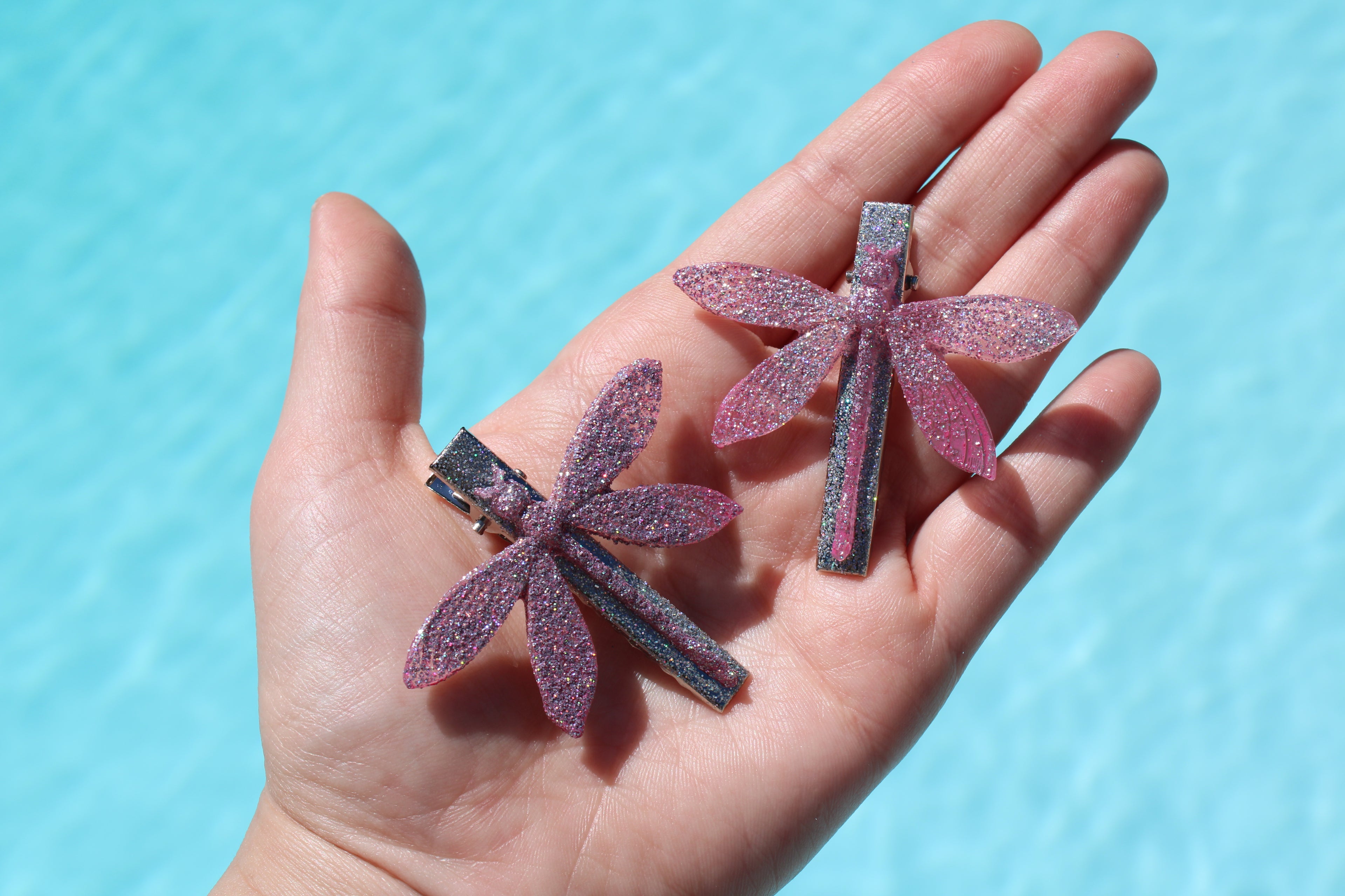 Hand holding two dragonfly-shaped hair clips with glittery finish against a blue background