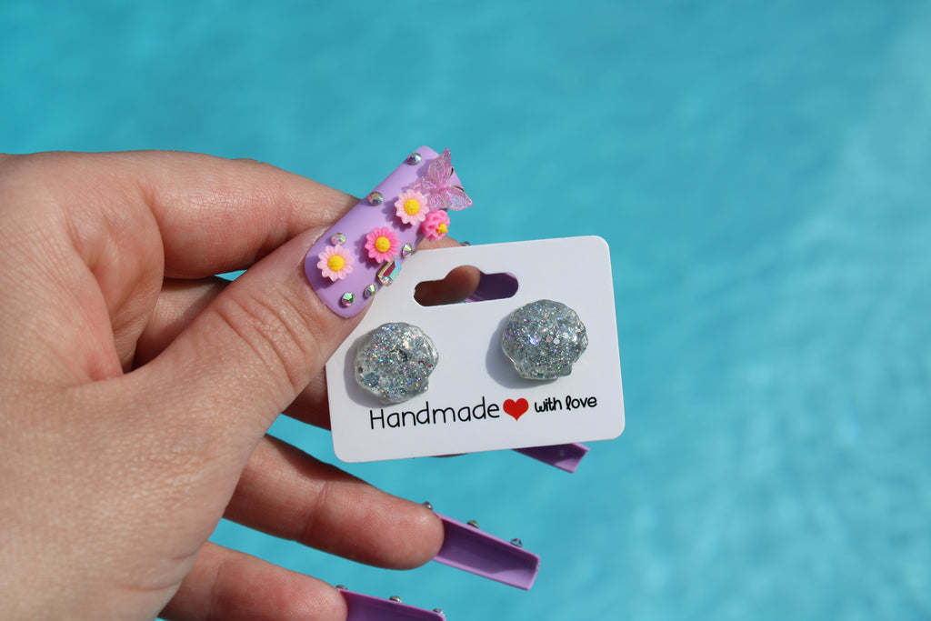 Handmade silver glitter seashell earrings on a card held by a hand with a pool background