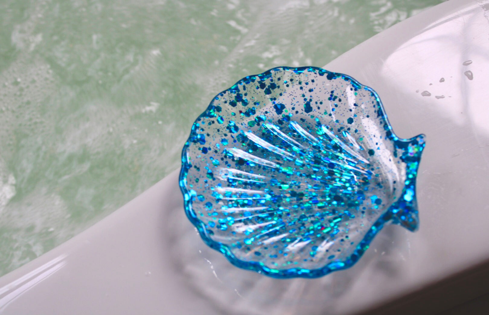 Blue glittery shell-shaped soap dish on a white surface with water in the background